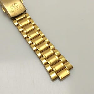 Puede incluir: Una correa de reloj dorada con un cierre con el logotipo de Seiko y la palabra "JAPAN". La correa está compuesta por eslabones rectangulares.