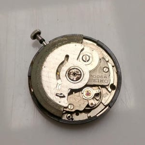 May include: A close-up of a Seiko 7006A watch movement. The mechanical components are visible, including gears, screws, and a balance wheel. The movement is circular, with a silver and gold color scheme. The text "SEIKO" is printed on the movement.