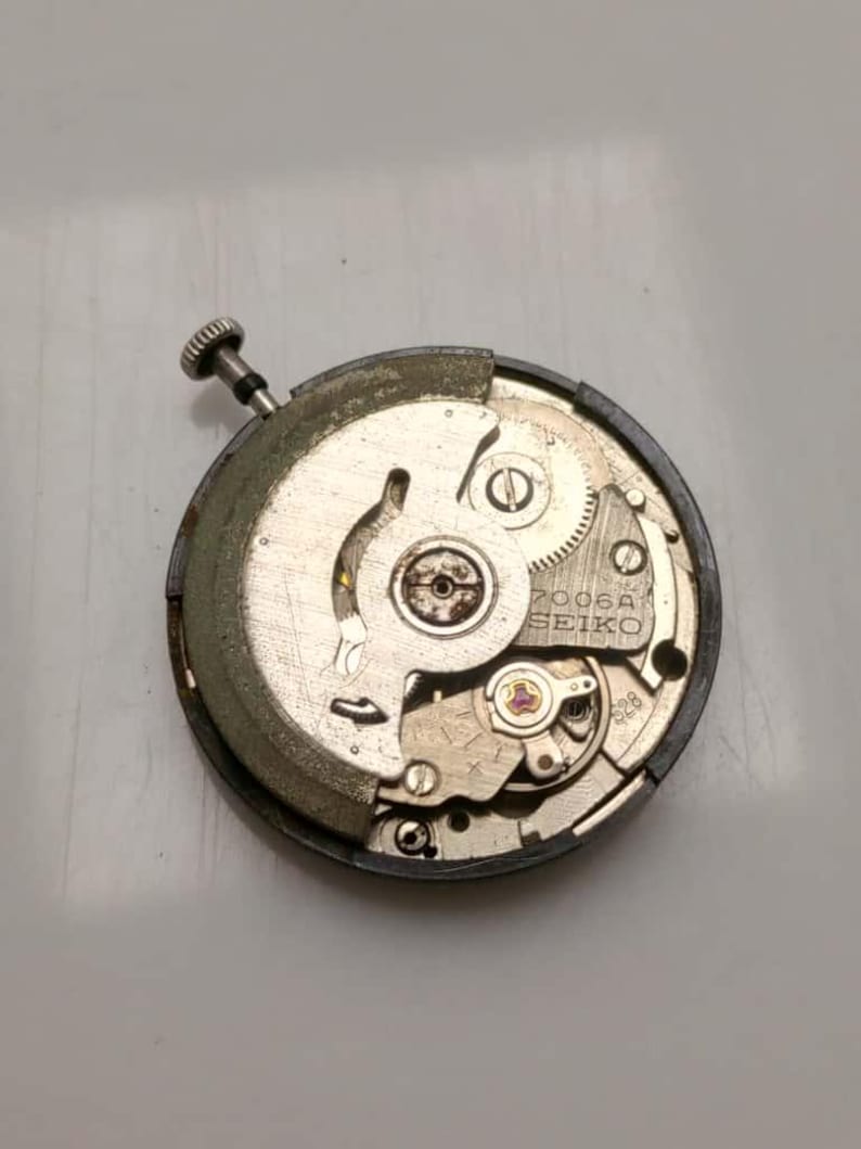 May include: A close-up of a Seiko 7006A watch movement. The mechanical components are visible, including gears, screws, and a balance wheel. The movement is circular, with a silver and gold color scheme. The text "SEIKO" is printed on the movement.