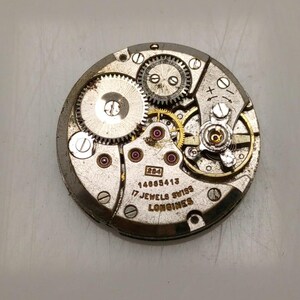May include: Close-up of a vintage watch movement with visible gears, jewels, and the text "17 Jewels Swiss Longines". The metal components are silver and gold-toned, with small red jewels. The watch mechanism is circular.