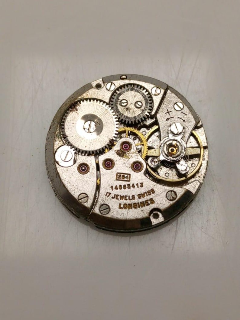 May include: Close-up of a vintage watch movement with visible gears, jewels, and the text "17 Jewels Swiss Longines". The metal components are silver and gold-toned, with small red jewels. The watch mechanism is circular.