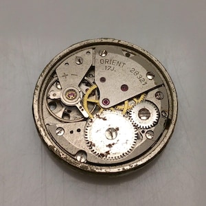 Puede incluir: Primer plano de un movimiento de reloj Orient vintage. El mecanismo del reloj presenta numerosos engranajes peque&ntilde;os, joyas y componentes met&aacute;licos. El texto "ORIENT 17J 28321" es visible en la esfera del reloj. El movimiento del reloj es circular y tiene un acabado plateado.