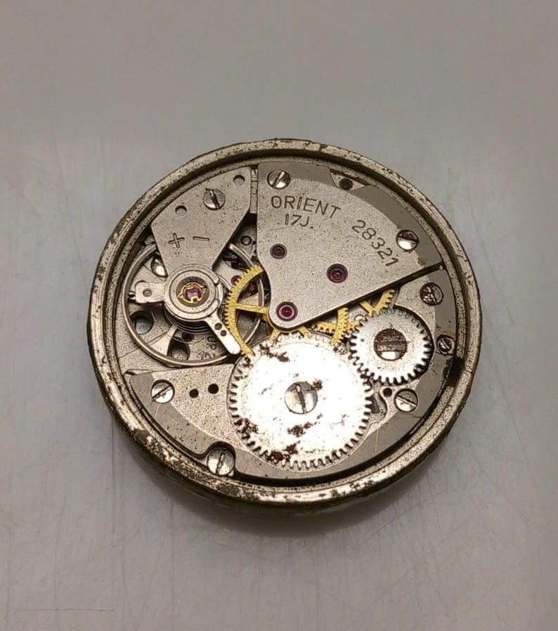 Puede incluir: Primer plano de un movimiento de reloj Orient vintage. El mecanismo del reloj presenta numerosos engranajes peque&ntilde;os, joyas y componentes met&aacute;licos. El texto "ORIENT 17J 28321" es visible en la esfera del reloj. El movimiento del reloj es circular y tiene un acabado plateado.