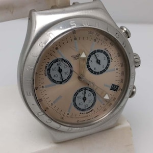 May include: A silver-toned Swatch watch with a light brown face. The watch features multiple dials for minutes, seconds, and decimals, with black numerals and hands. The watch has a date display and is marked "SWATCH AG 1999".