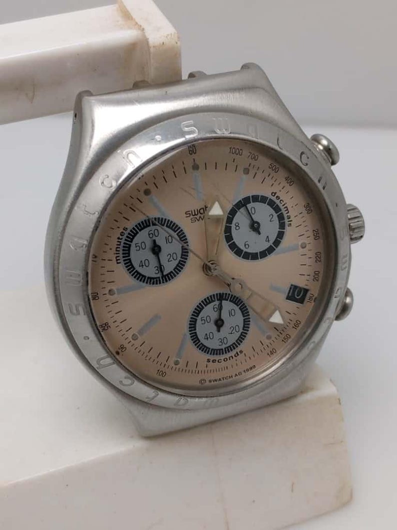 May include: A silver-toned Swatch watch with a light brown face. The watch features multiple dials for minutes, seconds, and decimals, with black numerals and hands. The watch has a date display and is marked "SWATCH AG 1999".