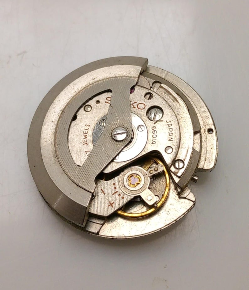 May include: A close-up of a Seiko watch movement, featuring intricate gears and components. The metal parts are silver and gold, with the text "SEIKO" and "JAPAN" visible. The movement is labeled "6601A" and "1 JEWELS".
