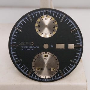 Op de afbeelding: Zwarte Seiko Chronograph Automatic wijzerplaat met zilveren sub-wijzerplaten en markeringen. Voorzien van de tekst "SEIKO CHRONOGRAPH AUTOMATIC" en "JAPAN G130". De wijzerplaat heeft een cirkelvormig ontwerp met een gedetailleerde minutenrail.