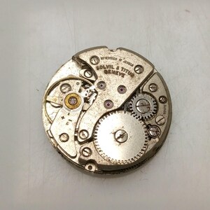 May include: Close-up of a vintage watch movement with the text "REVERTEEN 17 JEWELS SOLVIL & TITUS GENEVE". The silver-toned mechanism features gears, screws, and red jewels. The watch movement is circular and detailed.