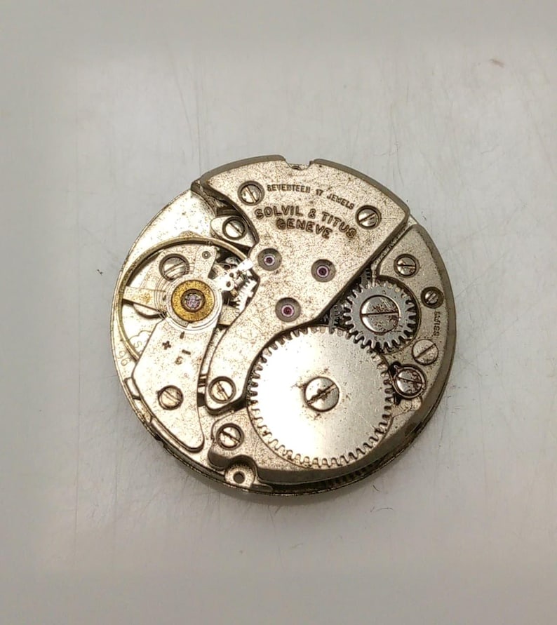 May include: Close-up of a vintage watch movement with the text "REVERTEEN 17 JEWELS SOLVIL & TITUS GENEVE". The silver-toned mechanism features gears, screws, and red jewels. The watch movement is circular and detailed.