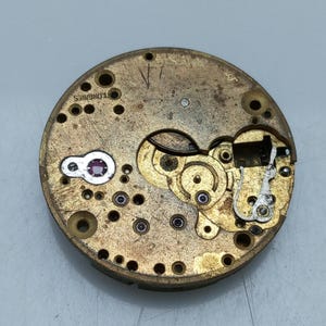 May include: Close-up of a vintage watch movement with intricate gears and components. The watch mechanism is circular, with a brassy-gold finish and visible text that reads "LONGINES". The design includes small jewels and various mechanical parts.