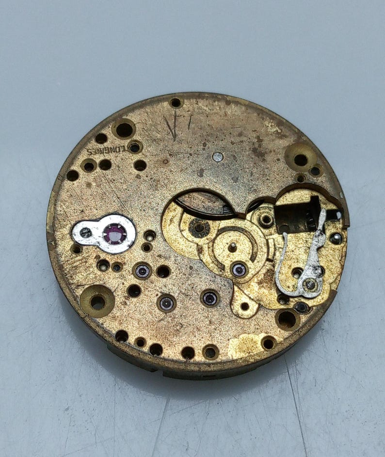 May include: Close-up of a vintage watch movement with intricate gears and components. The watch mechanism is circular, with a brassy-gold finish and visible text that reads "LONGINES". The design includes small jewels and various mechanical parts.