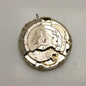 May include: A close-up of a watch movement with visible gears, screws, and a balance wheel. The metal components have a silver and gold color scheme. The watch movement is a mechanical component.