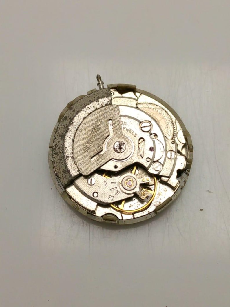 May include: A close-up of a watch movement with visible gears, screws, and a balance wheel. The metal components have a silver and gold color scheme. The watch movement is a mechanical component.