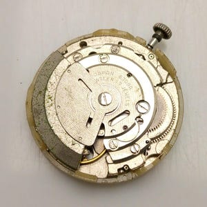 Może przedstawiać: Zdemontowany mechanizm zegarka z widocznymi kołami zębatymi, śrubami i wałem naciągowym. Srebrny mechanizm wyświetla napis "JAPAN SEVENTEEN 17 JEWELS 6309A". Zewnętrzna obudowa jest w jasnobeżowym kolorze.