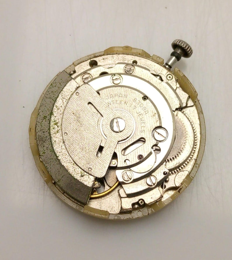 Może przedstawiać: Zdemontowany mechanizm zegarka z widocznymi kołami zębatymi, śrubami i wałem naciągowym. Srebrny mechanizm wyświetla napis "JAPAN SEVENTEEN 17 JEWELS 6309A". Zewnętrzna obudowa jest w jasnobeżowym kolorze.