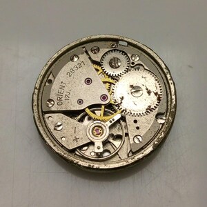Puede incluir: Primer plano de un movimiento de reloj Orient vintage. El mecanismo presenta numerosos engranajes peque&ntilde;os, tornillos y joyas. El texto "ORIENT 17J" y "28321" es visible en los componentes met&aacute;licos. El movimiento del reloj es circular y tiene una combinaci&oacute;n de colores plateado y dorado.
