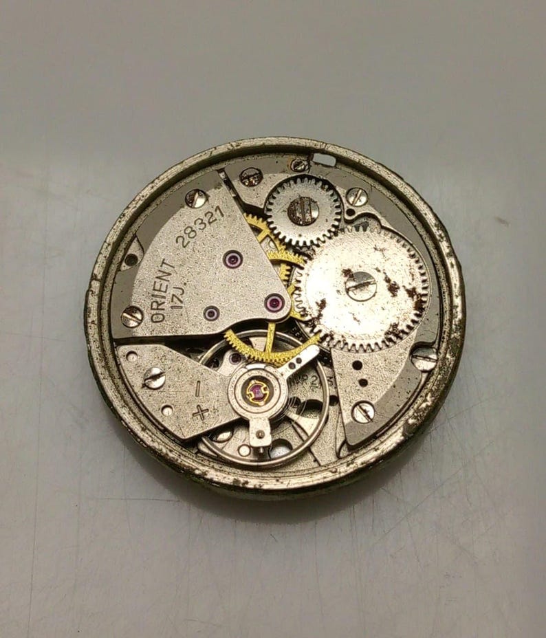 Puede incluir: Primer plano de un movimiento de reloj Orient vintage. El mecanismo presenta numerosos engranajes peque&ntilde;os, tornillos y joyas. El texto "ORIENT 17J" y "28321" es visible en los componentes met&aacute;licos. El movimiento del reloj es circular y tiene una combinaci&oacute;n de colores plateado y dorado.