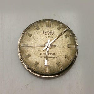 May include: A vintage gold-toned watch face, marked "ALSONS SEIKO" and "WATER PROOF". The round face features rectangular hour markers and silver hands. The watch face shows signs of age and wear.