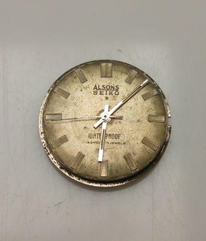 May include: A vintage gold-toned watch face, marked "ALSONS SEIKO" and "WATER PROOF". The round face features rectangular hour markers and silver hands. The watch face shows signs of age and wear.