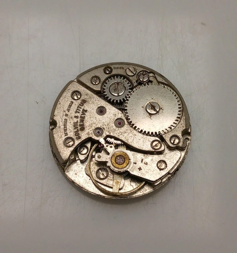 May include: Close-up of a vintage watch movement with gears, screws, and the text "SOLVIL & TITUS GENEVE". The watch mechanism is silver-toned with small red jewels and intricate details, showcasing the inner workings of a timepiece.