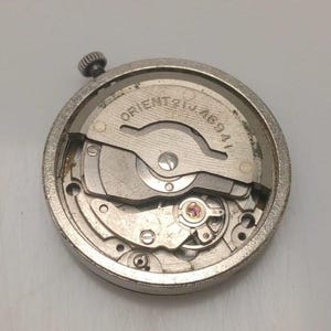 Può includere: Primo piano di un movimento di orologio argentato con la scritta "ORIENT 21J 46941". Il meccanismo comprende vari ingranaggi, molle e una piccola corona. Il movimento dell'orologio è circolare e sembra essere un pezzo vintage o di ricambio.