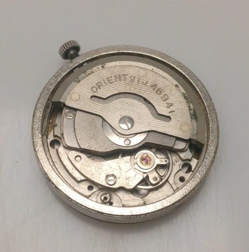 May include: A close-up of a silver-toned watch movement with the text "ORIENT 21J 46941". The watch mechanism features various gears, springs, and a small crown. The watch movement is circular and appears to be a vintage or replacement part.