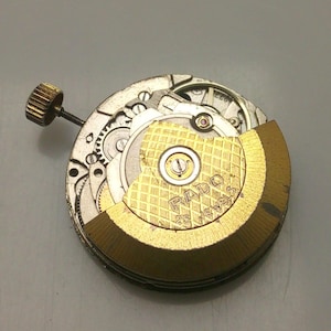May include: Close-up of a watch movement with visible gears, a gold-colored rotor, and the text "RADO 25 JEWELS". The watch mechanism has a gold-colored crown and is set against a gray background.