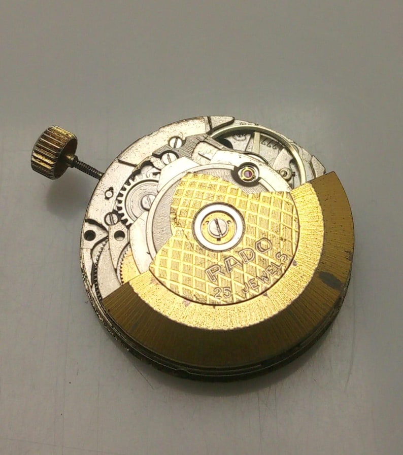 May include: Close-up of a watch movement with visible gears, a gold-colored rotor, and the text "RADO 25 JEWELS". The watch mechanism has a gold-colored crown and is set against a gray background.