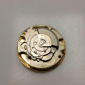 May include: Close-up of a vintage watch movement with intricate gears and components. The gold-toned metal mechanism features a circular design with various small parts, including a balance wheel and a Seiko label. The watch movement is a detailed example of horological engineering.