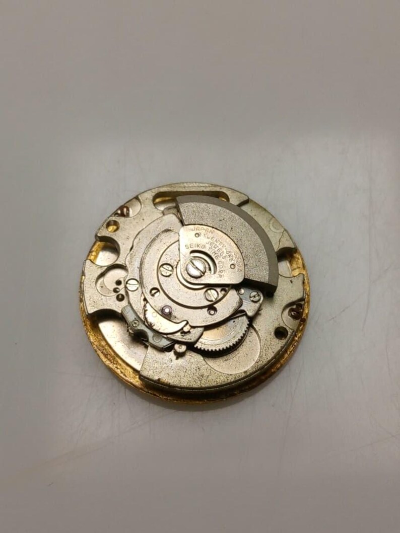 May include: Close-up of a vintage watch movement with intricate gears and components. The gold-toned metal mechanism features a circular design with various small parts, including a balance wheel and a Seiko label. The watch movement is a detailed example of horological engineering.