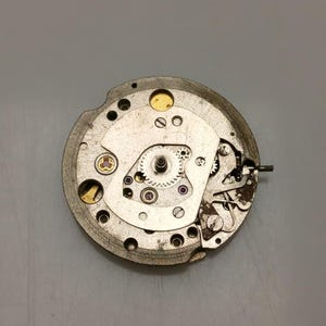 May include: Close-up of a mechanical watch movement. The silver-colored mechanism features various gears, screws, and jewels. The intricate design showcases the internal components of a timepiece, highlighting its precision and craftsmanship.