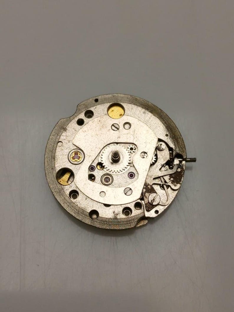 May include: Close-up of a mechanical watch movement. The silver-colored mechanism features various gears, screws, and jewels. The intricate design showcases the internal components of a timepiece, highlighting its precision and craftsmanship.