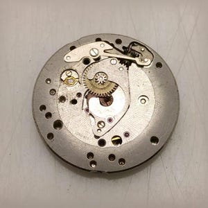 May include: Close-up of a vintage watch movement with intricate gears, screws, and jewels. The silver-toned metal components are arranged in a circular pattern, showcasing the mechanical complexity of the timepiece. The word "LONGINES" is visible.