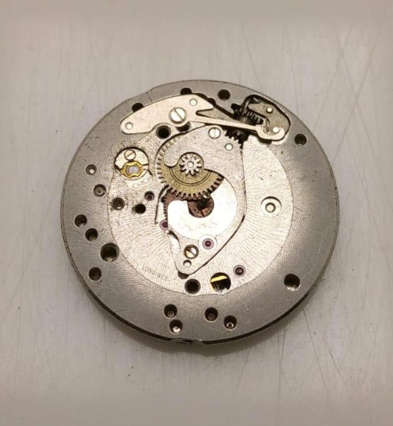 May include: Close-up of a vintage watch movement with intricate gears, screws, and jewels. The silver-toned metal components are arranged in a circular pattern, showcasing the mechanical complexity of the timepiece. The word "LONGINES" is visible.