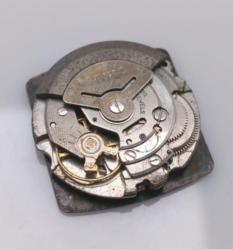 Puede incluir: Primer plano de un movimiento de reloj vintage, mostrando engranajes, resortes y la marca "SEIKO". Los componentes met&aacute;licos son plateados y dorados, revelando los detalles internos del reloj.