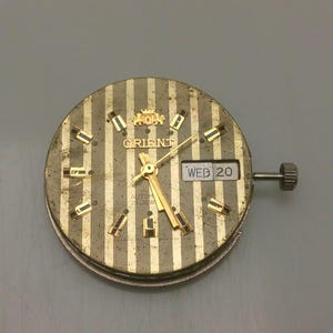 Pode incluir: Um mostrador de rel&oacute;gio Orient vintage com um design listrado dourado e prateado. O mostrador apresenta ponteiros, marcadores e a exibi&ccedil;&atilde;o do dia e da data dourados. O texto "ORIENT" &eacute; vis&iacute;vel na parte superior e a data &eacute; "QUA 20".
