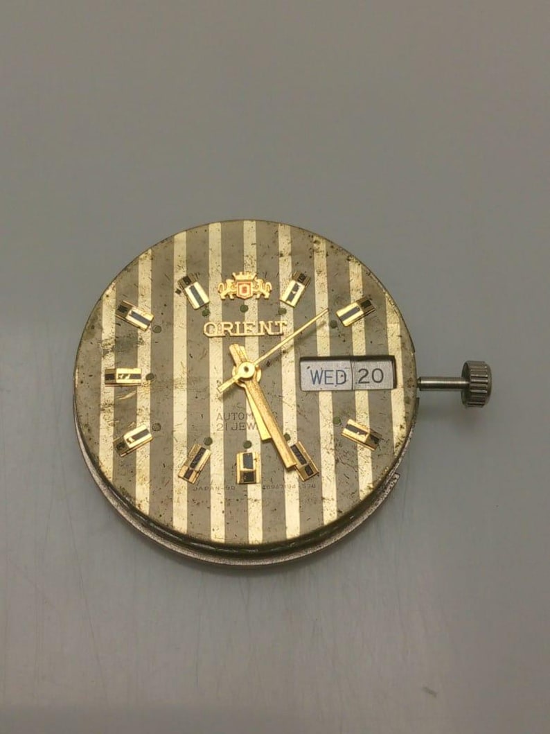 Pode incluir: Um mostrador de rel&oacute;gio Orient vintage com um design listrado dourado e prateado. O mostrador apresenta ponteiros, marcadores e a exibi&ccedil;&atilde;o do dia e da data dourados. O texto "ORIENT" &eacute; vis&iacute;vel na parte superior e a data &eacute; "QUA 20".