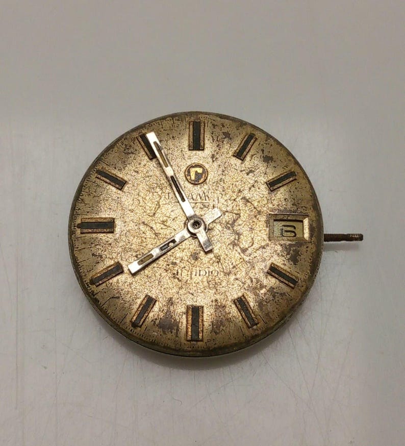 May include: A vintage watch face with a gold-toned, textured surface. The face features black rectangular hour markers, gold hands, and a date window displaying the number 6. The watch face has the brand name "AVIA" and "IDIO" printed on it.