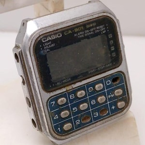 May include: Vintage Casio CA-901 digital watch with calculator function. The watch has a silver-tone case, a dark display, and blue buttons. Features include light, adjust, and calculator functions. The watch face displays the text "CASIO CA-901".