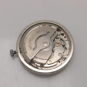 May include: A close-up of a silver watch movement with visible gears, springs, and a winding stem. The watch mechanism has the text "ORIENT 21 JEWELS 46941" engraved on the metal. The watch movement is circular and has a brushed metal finish.