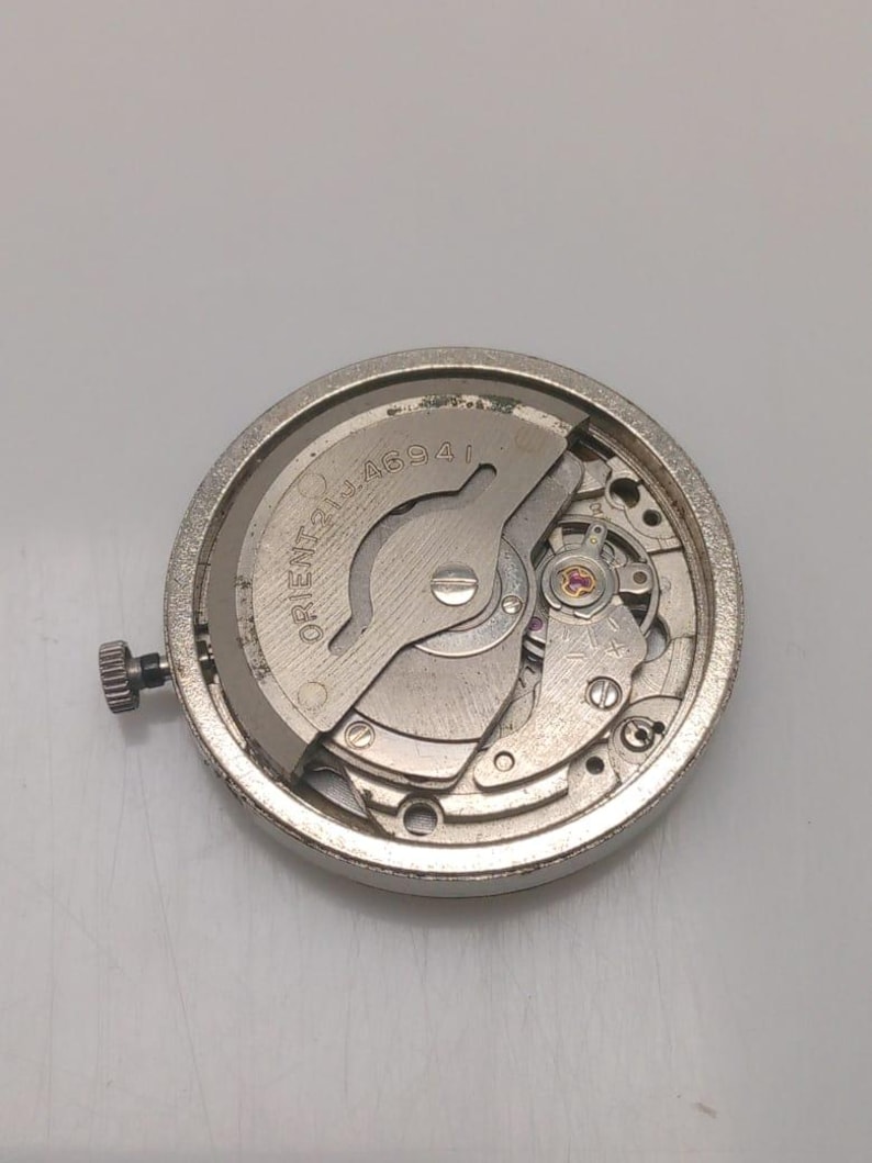 May include: A close-up of a silver watch movement with visible gears, springs, and a winding stem. The watch mechanism has the text "ORIENT 21 JEWELS 46941" engraved on the metal. The watch movement is circular and has a brushed metal finish.