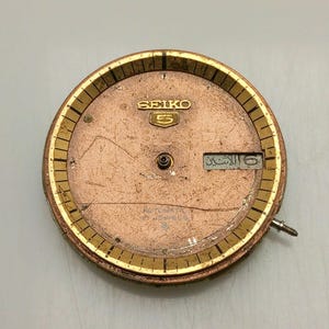 May include: A vintage Seiko 5 watch face with a copper-colored dial and gold-toned bezel. The dial features the Seiko logo and a day-date window. The watch face has a screw and is likely a replacement part.