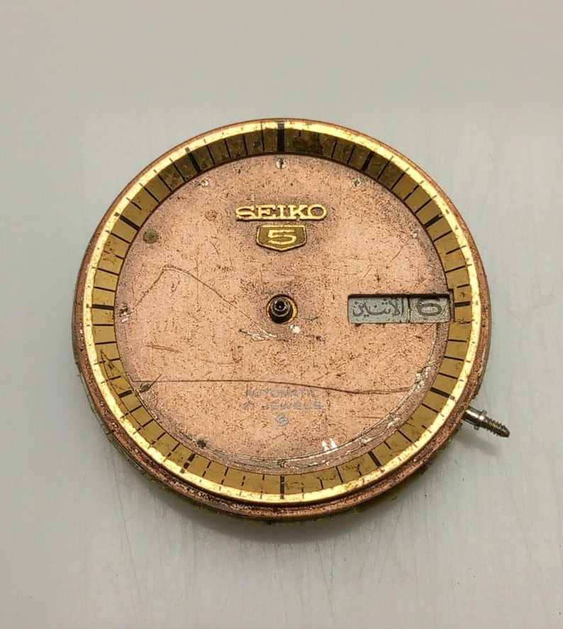 May include: A vintage Seiko 5 watch face with a copper-colored dial and gold-toned bezel. The dial features the Seiko logo and a day-date window. The watch face has a screw and is likely a replacement part.