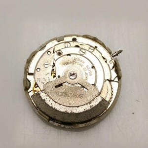 May include: A close-up of a Seiko watch movement, featuring intricate gears and components. The watch mechanism is silver and gold-toned, with the text "SEIKO" and "JAPAN TWENTYONE 21 JEWELS" visible. The watch movement is on a white surface.