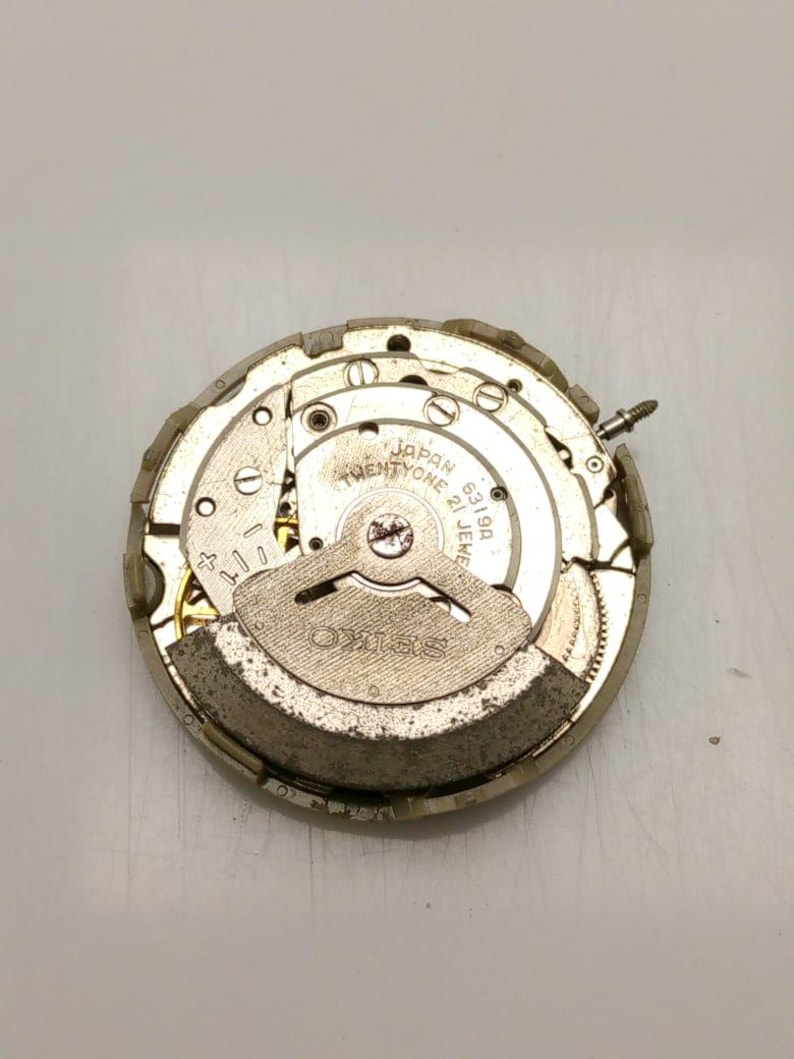 May include: A close-up of a Seiko watch movement, featuring intricate gears and components. The watch mechanism is silver and gold-toned, with the text "SEIKO" and "JAPAN TWENTYONE 21 JEWELS" visible. The watch movement is on a white surface.