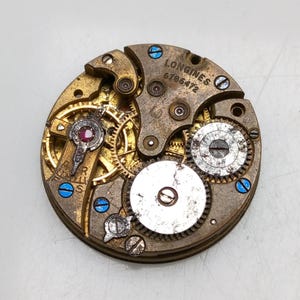 May include: Close-up of a vintage Longines watch movement. The mechanism features intricate gears, a ruby jewel, and the text "LONGINES 6786472". The metal components are gold and silver with blue accents.
