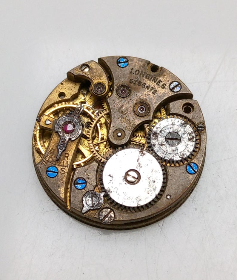 May include: Close-up of a vintage Longines watch movement. The mechanism features intricate gears, a ruby jewel, and the text "LONGINES 6786472". The metal components are gold and silver with blue accents.
