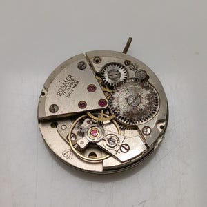 May include: Close-up of a disassembled Roamer watch movement. The silver-toned mechanism features various gears, jewels, and the text "Roamer 17 Jewels Swiss Made". The watch movement is circular and shows signs of age.