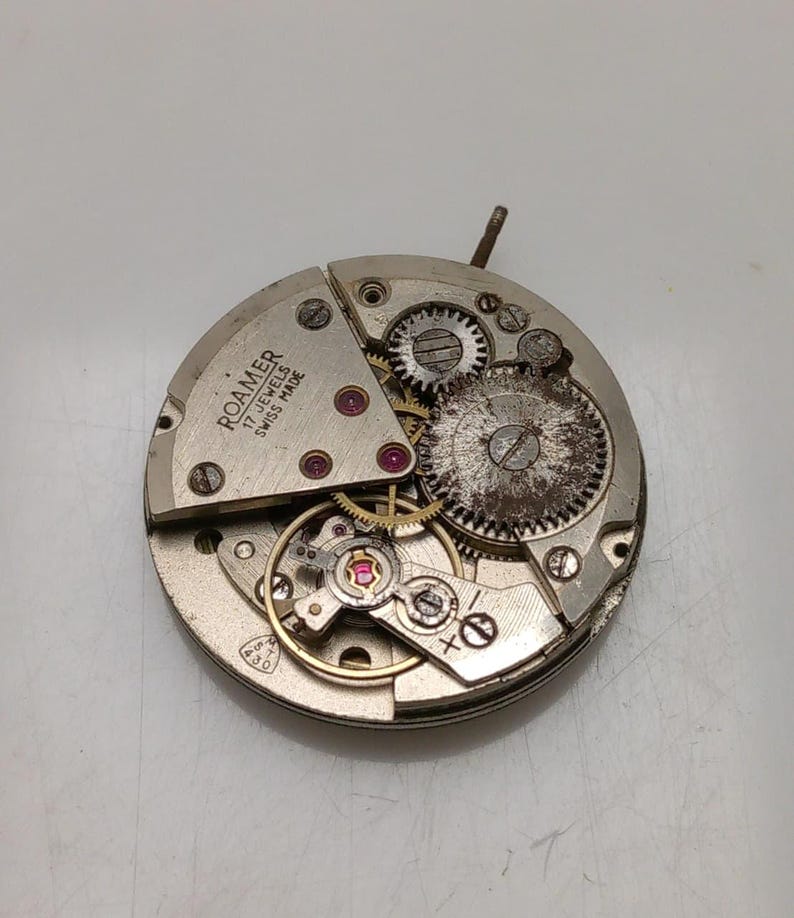 May include: Close-up of a disassembled Roamer watch movement. The silver-toned mechanism features various gears, jewels, and the text "Roamer 17 Jewels Swiss Made". The watch movement is circular and shows signs of age.