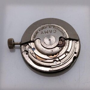 May include: A close-up of a watch movement with the brand name "CAMY" visible. The movement is silver-toned with intricate gears, screws, and a winding stem. The text "TWENTY-FIVE JEWELS SWISS" is also present.